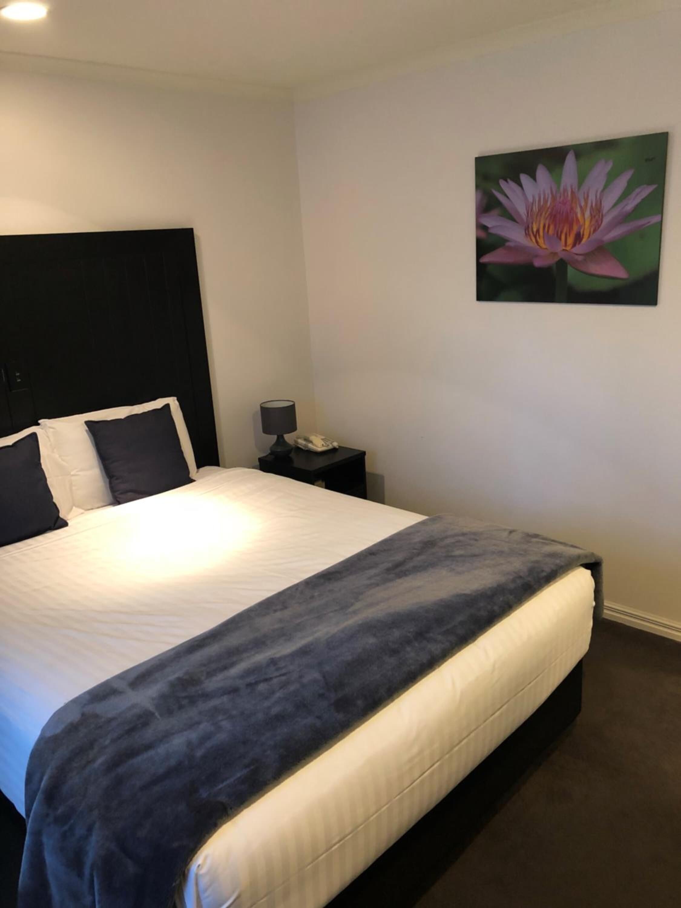 A hotel room with a bed, a nightstand, a lamp, and a wall-mounted painting of a flower.
