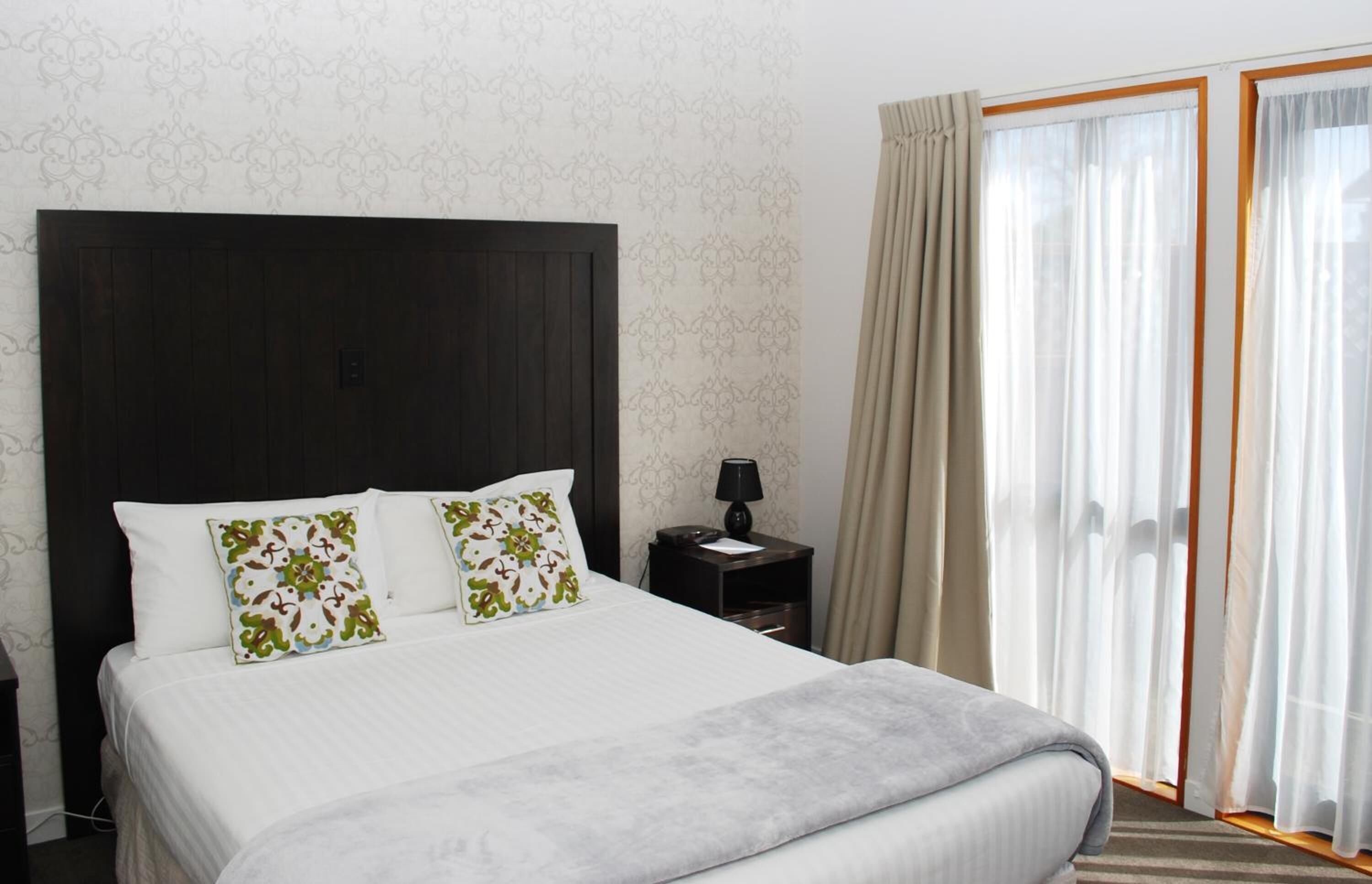 Camera d'albergo con un letto grande, un comodino, una lampada e vista all'esterno attraverso tende leggere.