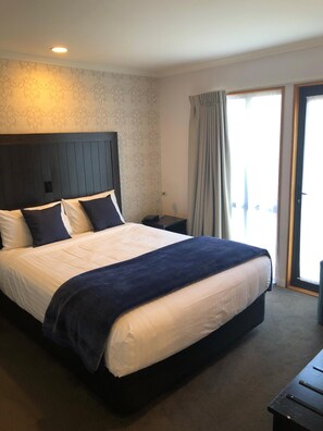 A hotel room with a large bed, a nightstand, a lamp, and a view of the outdoors through a window with curtains.
