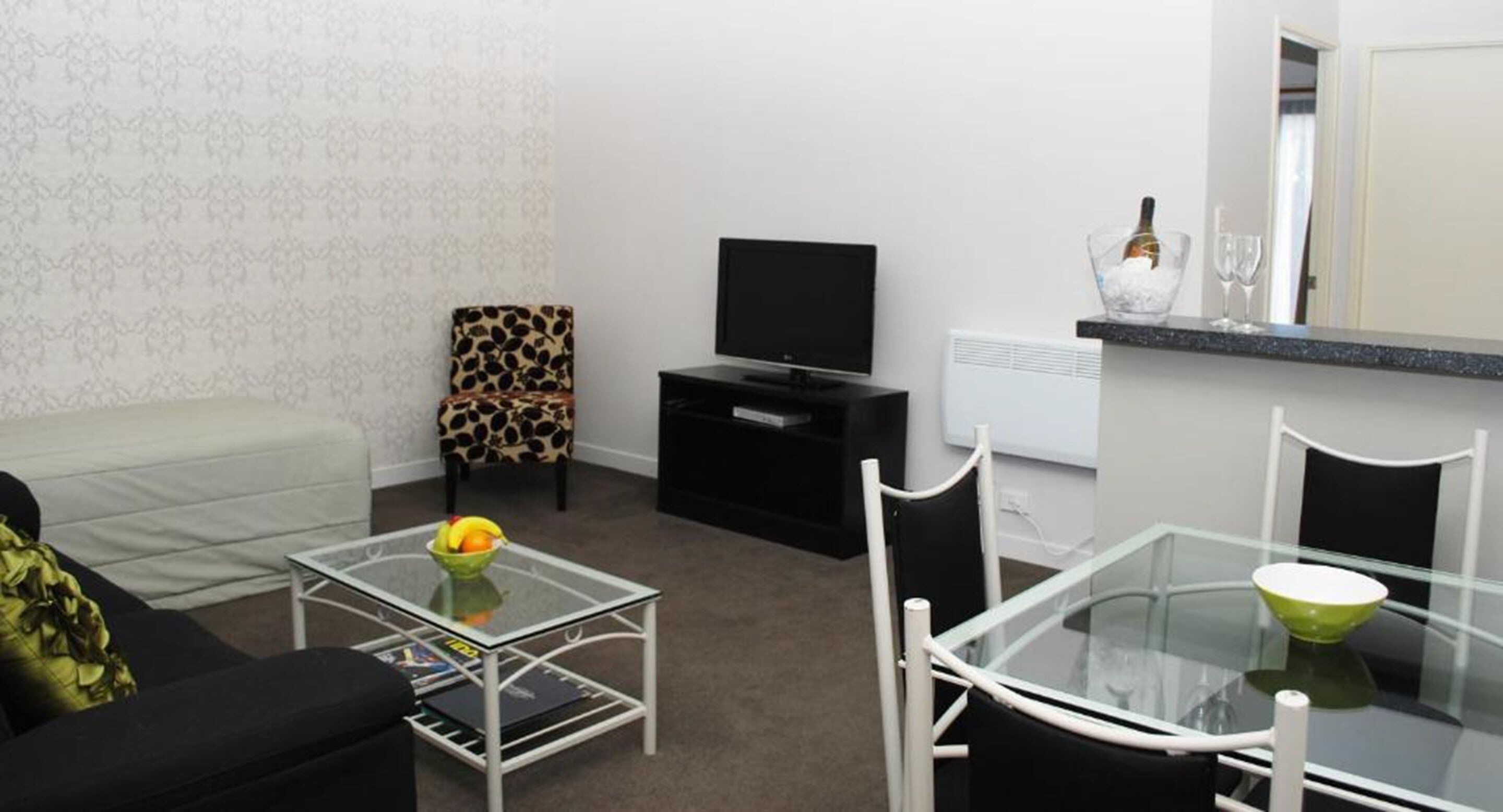 Una sala moderna con sofá, una mesa de centro de vidrio con fruta, televisor, una pequeña mesa de comedor con sillas y una pared blanca con papel tapiz estampado.