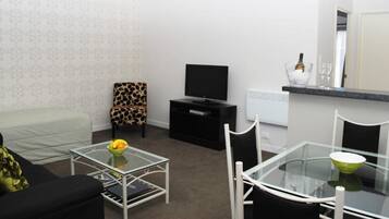 A modern living room with a sofa, a glass coffee table with fruit, a TV, a small dining table with chairs, and a white wall with a patterned wallpaper.