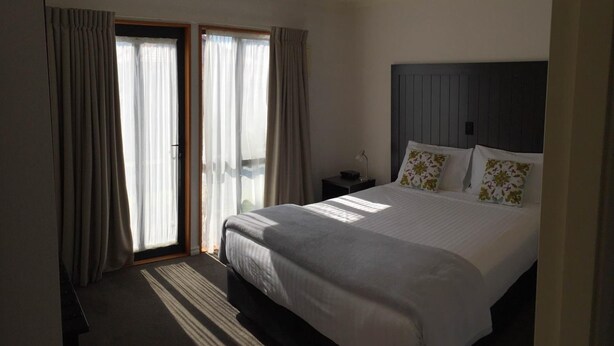 A hotel room with a bed, a nightstand, a lamp, and a view through a window with curtains. - Bethlehem Motor Inn (Tauranga)