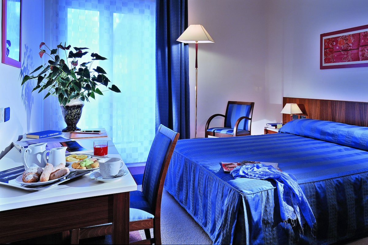 A hotel room with a large bed, a desk with breakfast items, a chair, and a bedside table with a lamp.