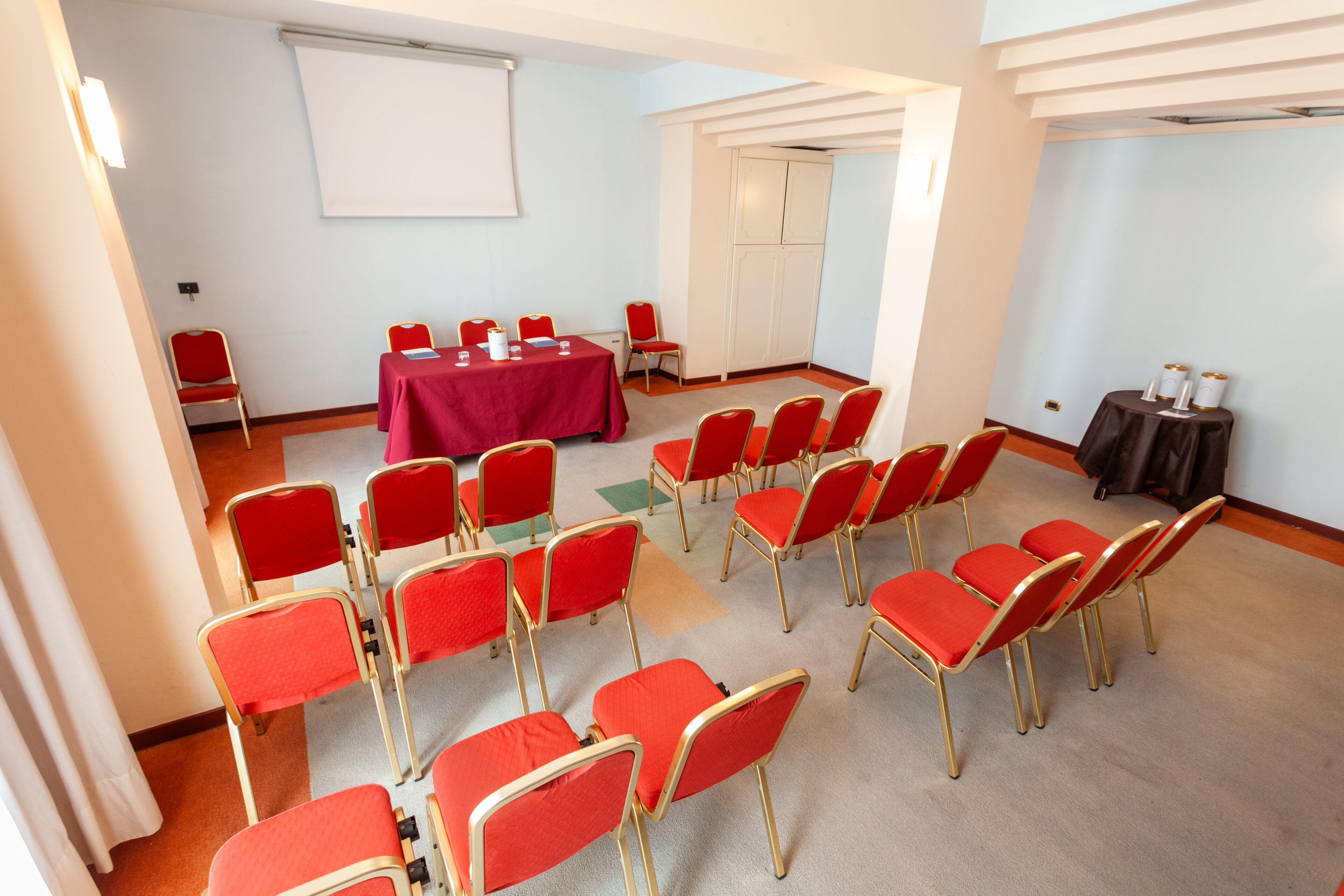 A conference room with rows of red chairs, a table with a red tablecloth, and a projector screen.