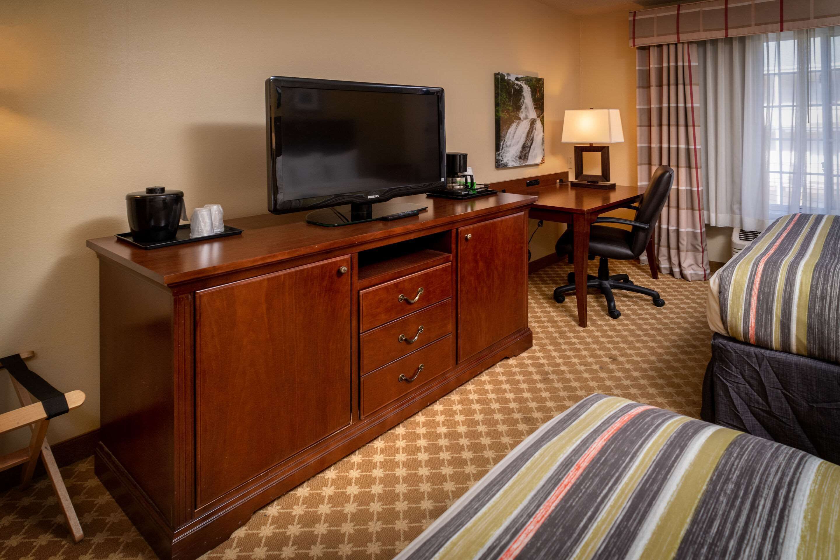 Una habitación de hotel con un escritorio de madera, una televisión, una cama con ropa de cama a rayas, una silla y un cuadro de una cascada en la pared.