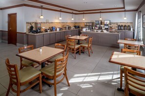 A hotel dining area with multiple tables and chairs, a kitchen area with various appliances and food items, and a door leading to another room.