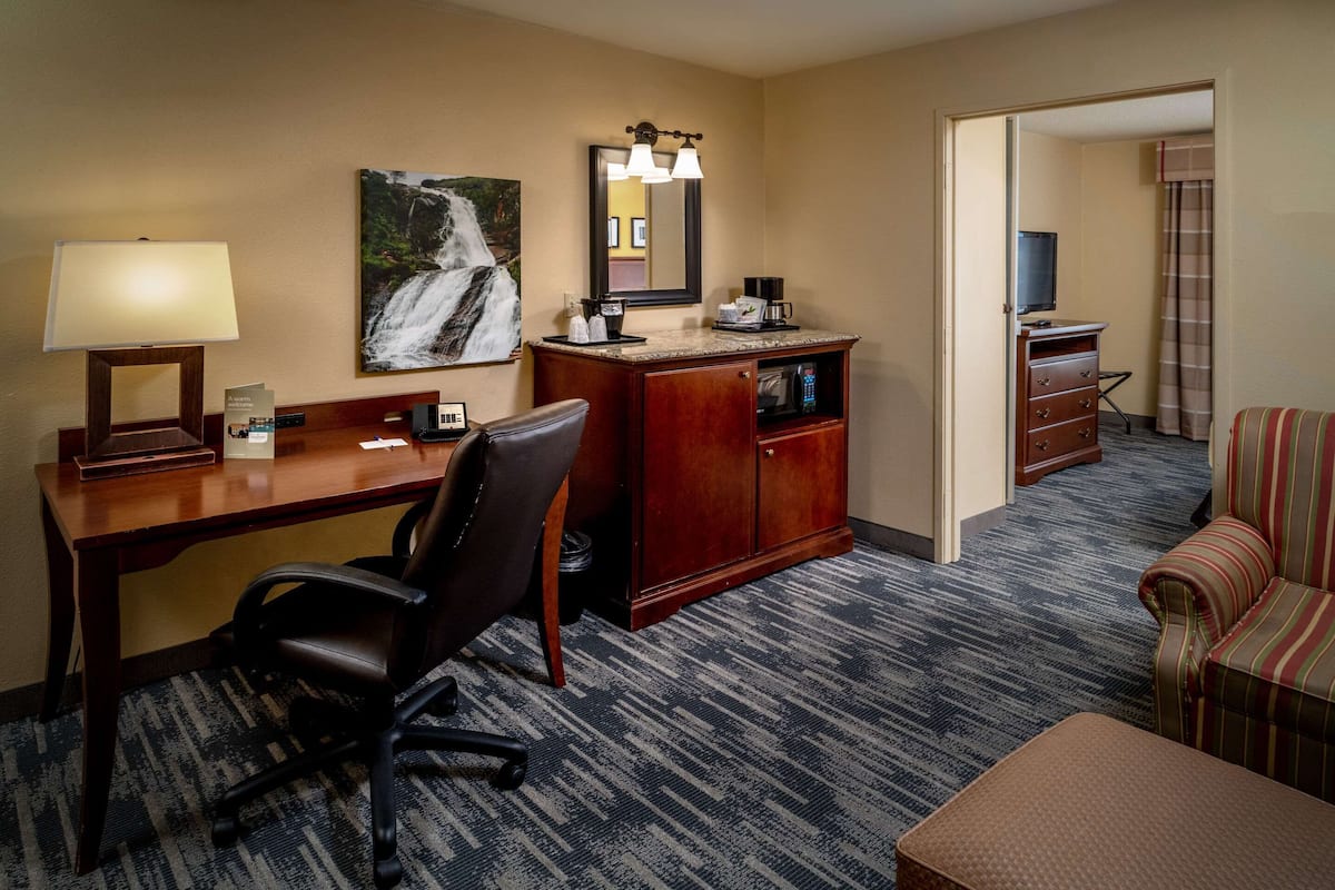 A hotel room with a desk, office chair, and a large mirror above it. There is a bed with striped bedding and a small table with a lamp.