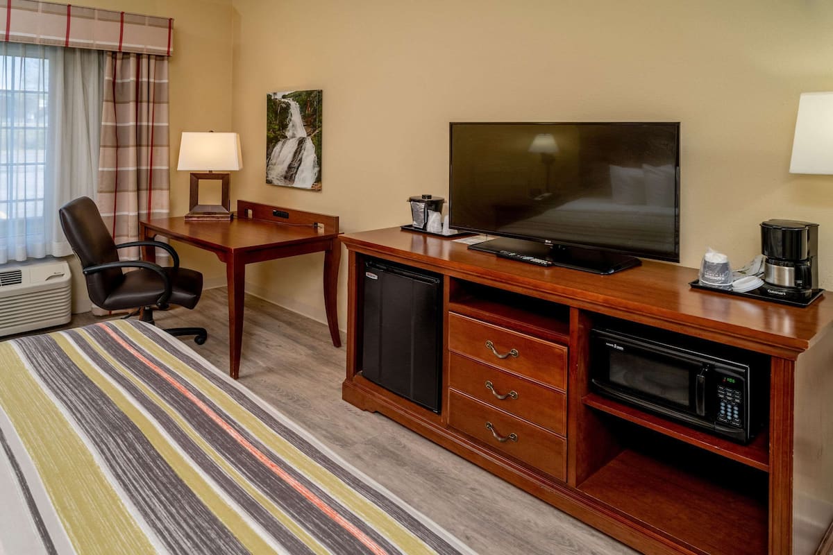 A hotel room with a wooden desk, a television, a microwave, a coffee maker, a chair, a lamp, and a painting on the wall.