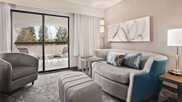 A modern hotel room with a grey sofa, a matching armchair, a small table, and a large window with a view of trees. - Courtyard by Marriott San Jose South/Morgan Hill (Morgan Hill)