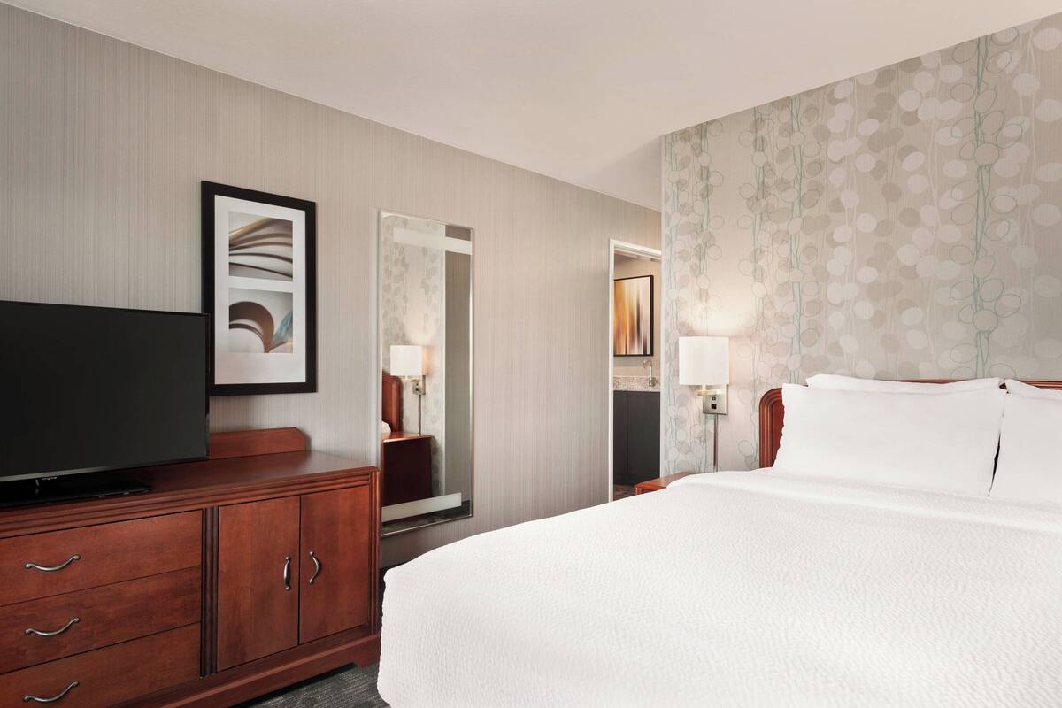 Une chambre d’hôtel avec un grand lit, un téléviseur à écran plat posé sur une commode en bois, un miroir et un papier peint à motifs floraux.