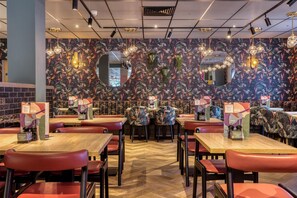 L’intérieur d’un restaurant avec des tables en bois, des chaises rouges, un papier peint à motifs floraux et des lampes suspendues.