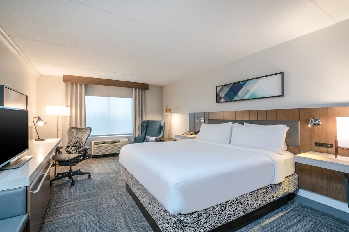A hotel room with a large bed, a desk with a computer, a chair, a TV, and a window with curtains.