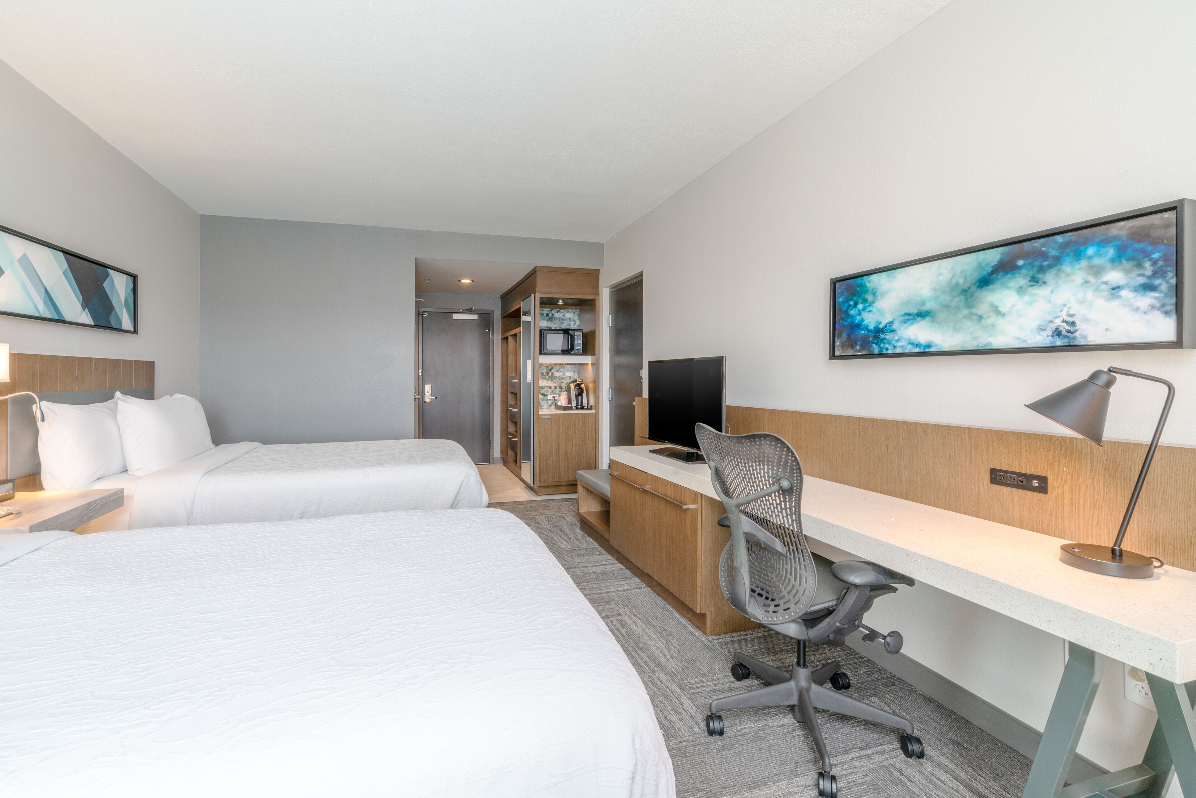A hotel room with two beds, a desk, a chair, a TV, and a microwave.