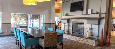A spacious dining area with a long wooden table and blue upholstered chairs, a fireplace with a stone surround, and a flat-screen TV mounted above it.