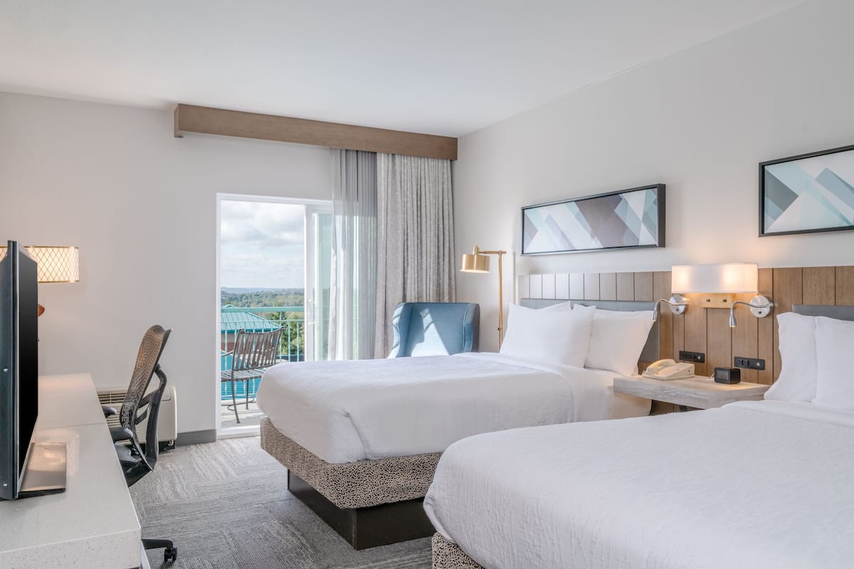 A hotel room with two beds, a desk, a chair, a TV, and a balcony with a view.