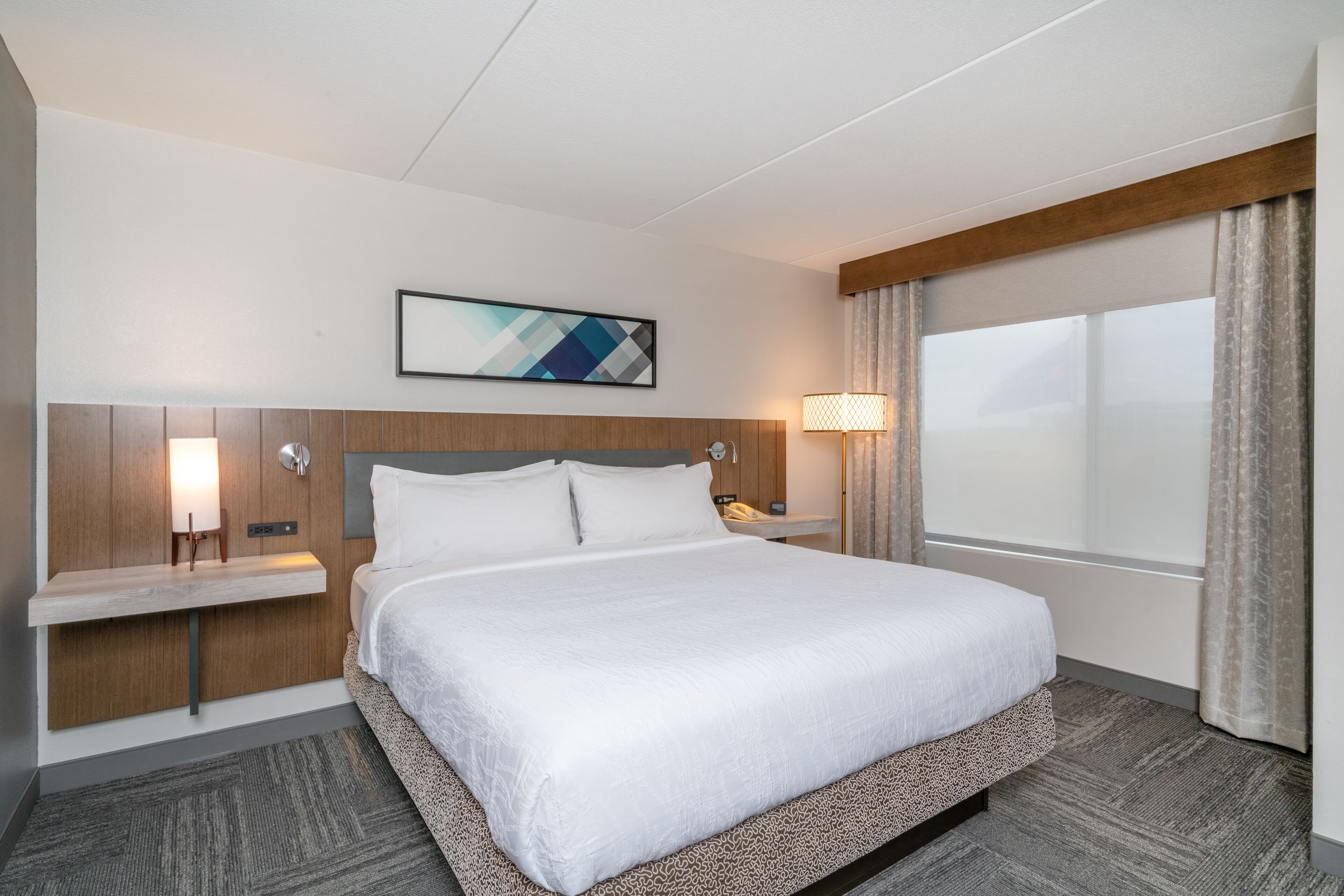 Camera d'albergo con un letto grande, una testiera in legno, un comodino con una lampada, un'opera d'arte appesa al muro e una finestra con tende.