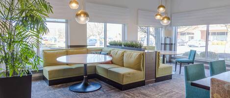 Un lobby moderno con sofá modular, una mesa redonda y sillas. Hay una planta grande en maceta y vistas al exterior a través de amplias ventanas.