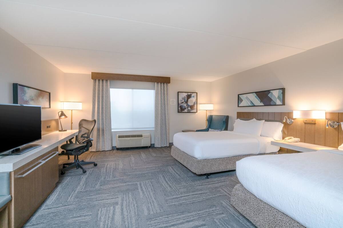 A hotel room with a bed, a desk with a TV, a chair, a lamp, and a window with curtains.