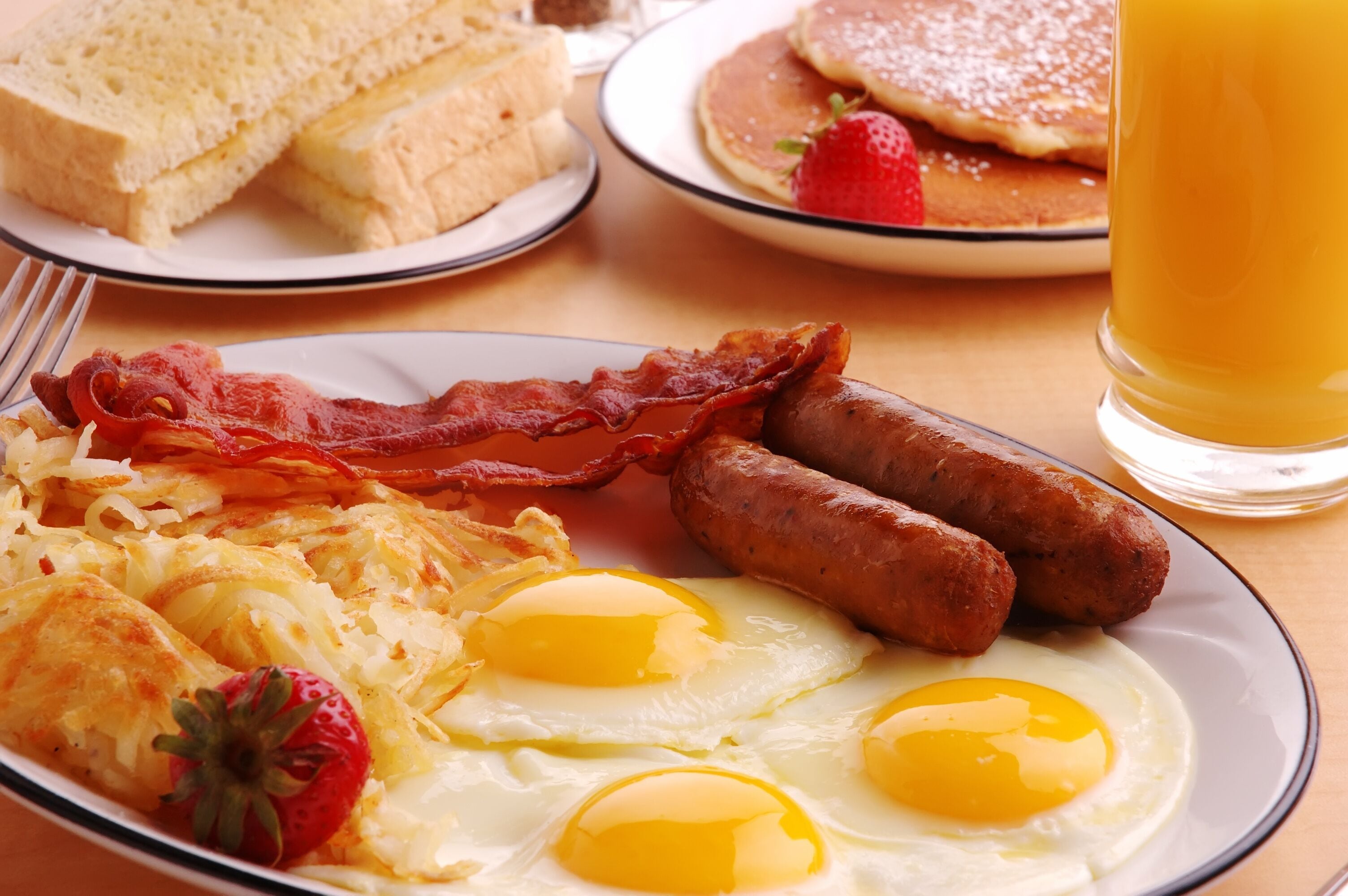 Un plato de desayuno con tocino, salchichas, huevos, tostadas, panqueques y un vaso de jugo de naranja.