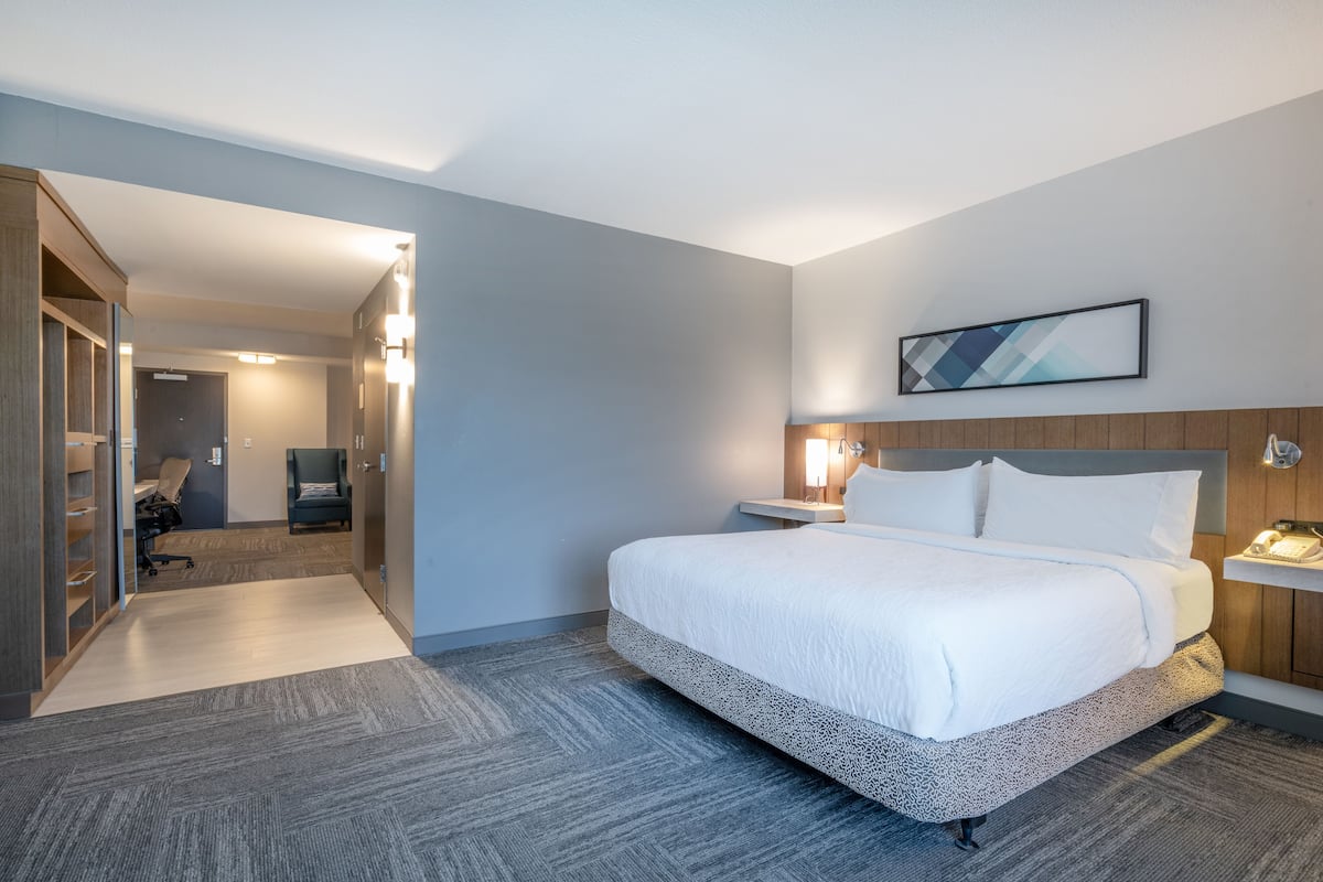 A hotel room with a large bed, a bedside table with a lamp, a wall-mounted phone, and a door leading to a hallway with a desk and chair.
