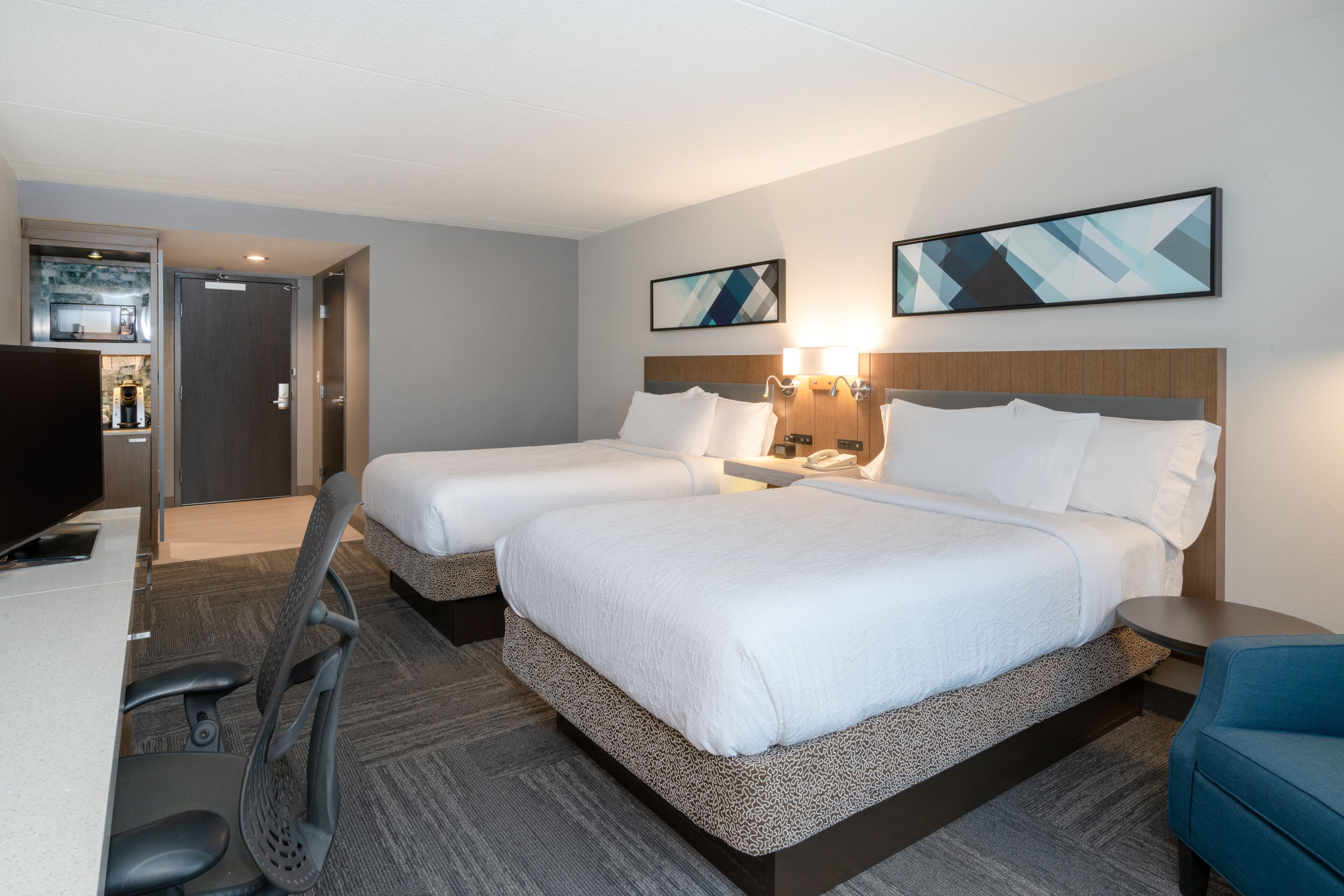 A hotel room with two beds, a desk, a chair, and a TV.