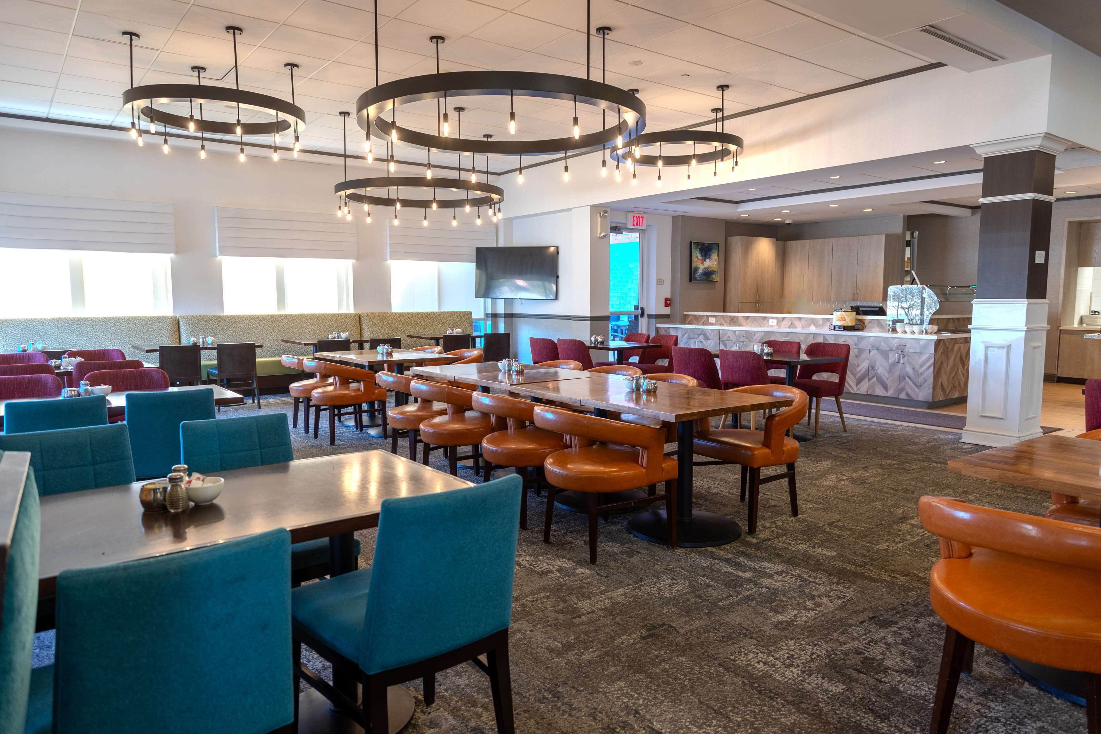 A modern hotel lobby with multiple dining areas featuring round tables and chairs, a central bar area, and a large screen TV.