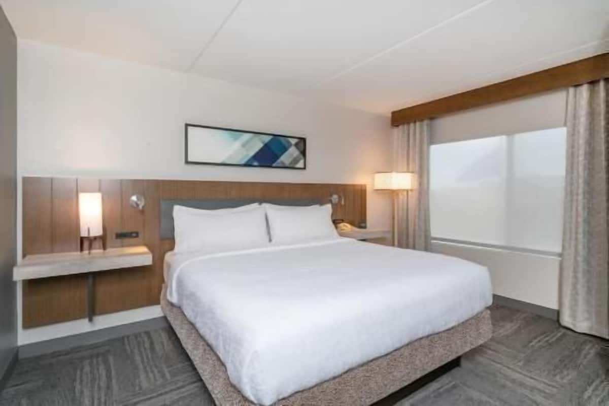 A hotel room with a bed, a nightstand, a lamp, a painting on the wall, and a window with curtains.