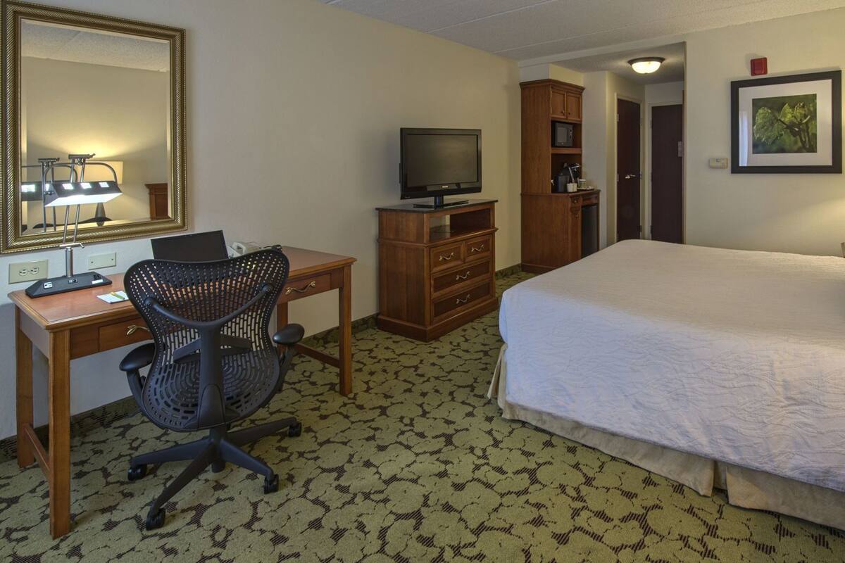 A hotel room with a bed, desk, chair, television, wardrobe, and a mirror.