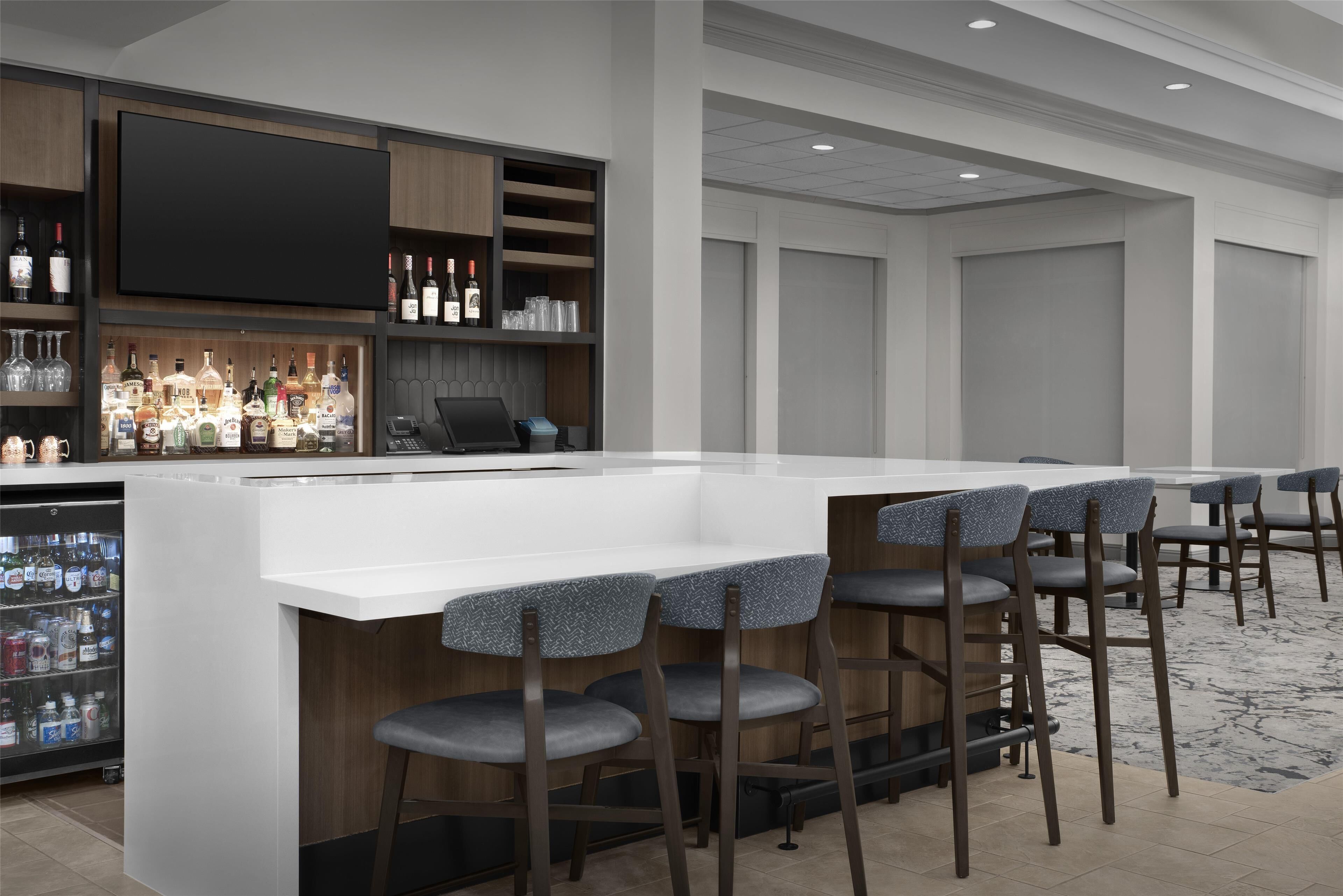 A bar area with a white countertop, multiple bar stools, a television, and shelves stocked with bottles and glasses.