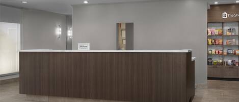 Zona de recepción de hotel con un mostrador de madera, un letrero que dice 'Hilton' y una tienda llamada 'The Shop' al fondo.