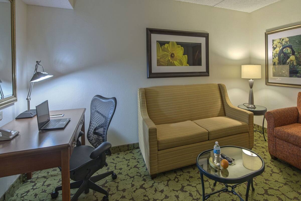 A hotel room with a sofa, armchair, desk with laptop, chair, and two framed pictures on the wall.