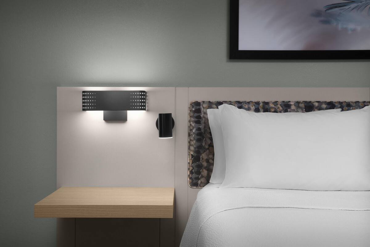 Une chambre d’hôtel bien rangée, avec un lit, une table de chevet et un luminaire fixé au mur.