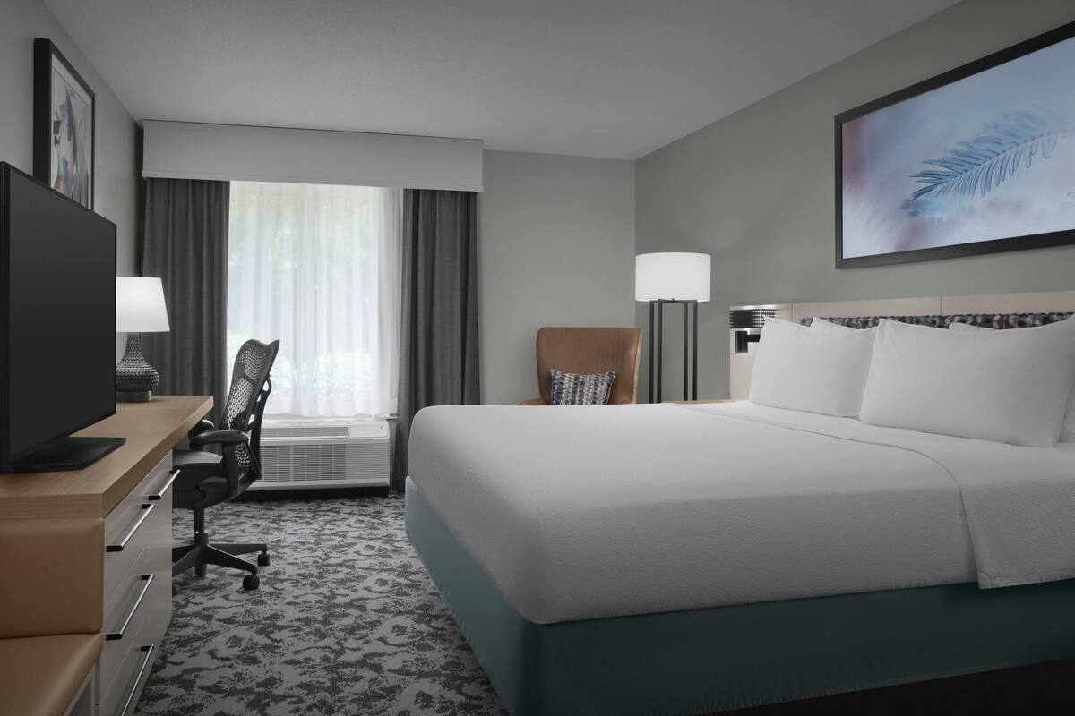 Une chambre d’hôtel avec un grand lit, un bureau avec un ordinateur, une chaise, une lampe de bureau et une grande fenêtre avec des rideaux.