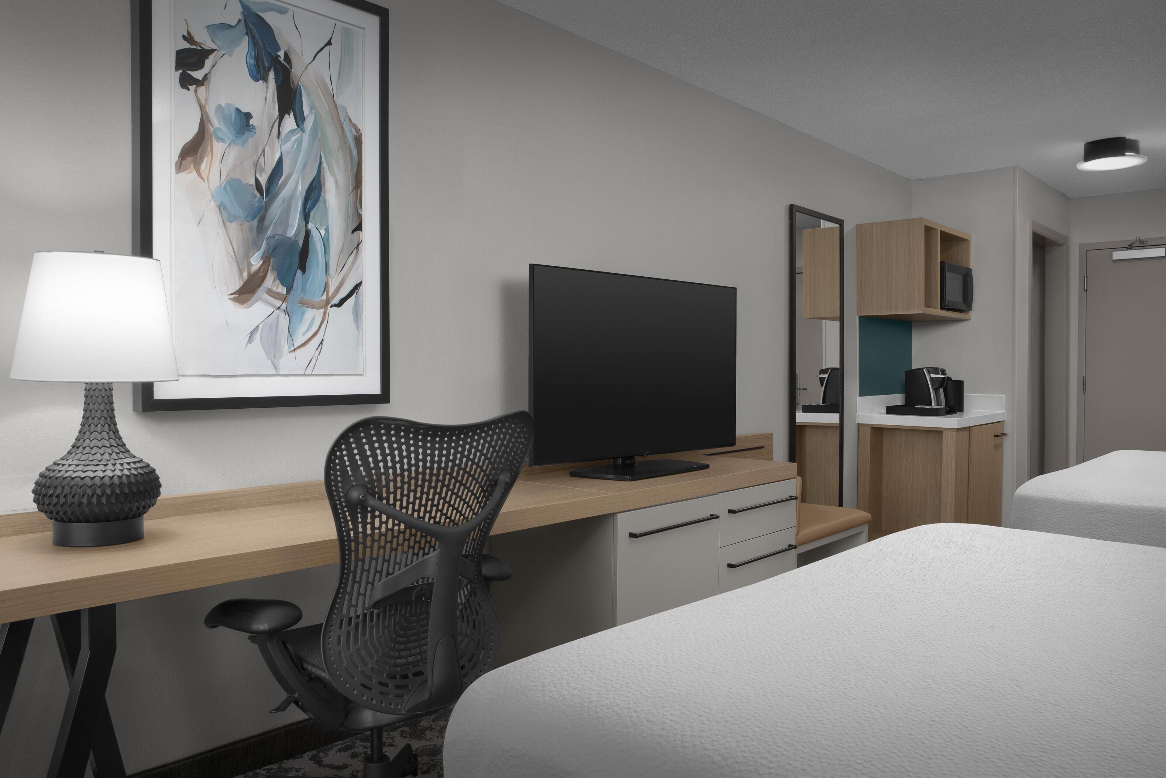 A modern hotel room with a bed, a desk with a computer, a chair, a TV, a lamp, a mirror, and a microwave.