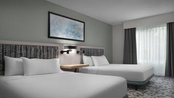 Habitación de hotel con dos camas, cabecera con una obra de arte de paisaje urbano, mesita de noche con lámpara y un ventanal con cortinas.