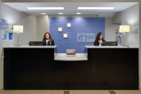 Bancada de recepção com dois funcionários, parede azul com o logotipo e um livro pequeno no balcão.