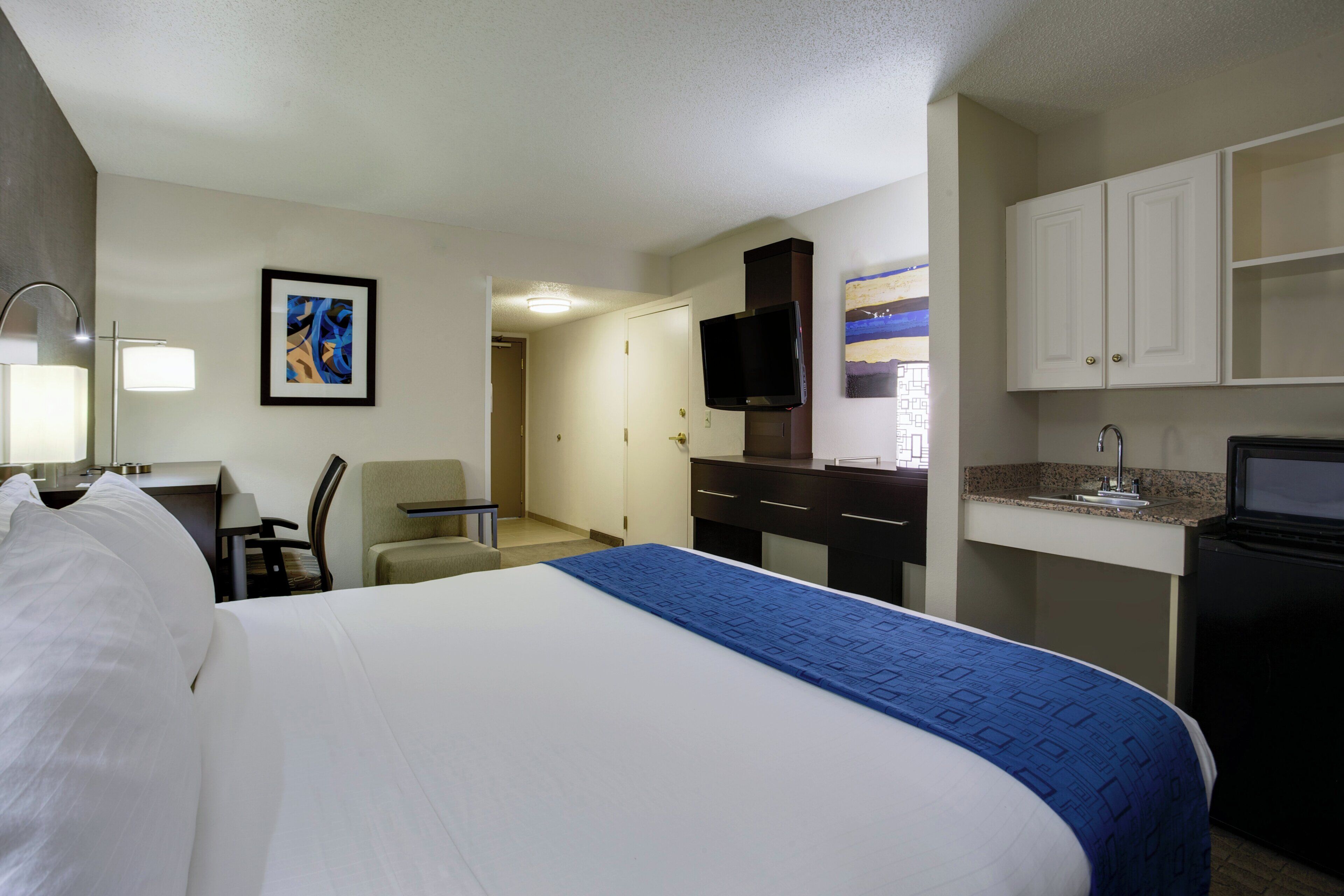 A hotel room with a bed, a desk, a chair, a television, a kitchenette, and a painting on the wall.