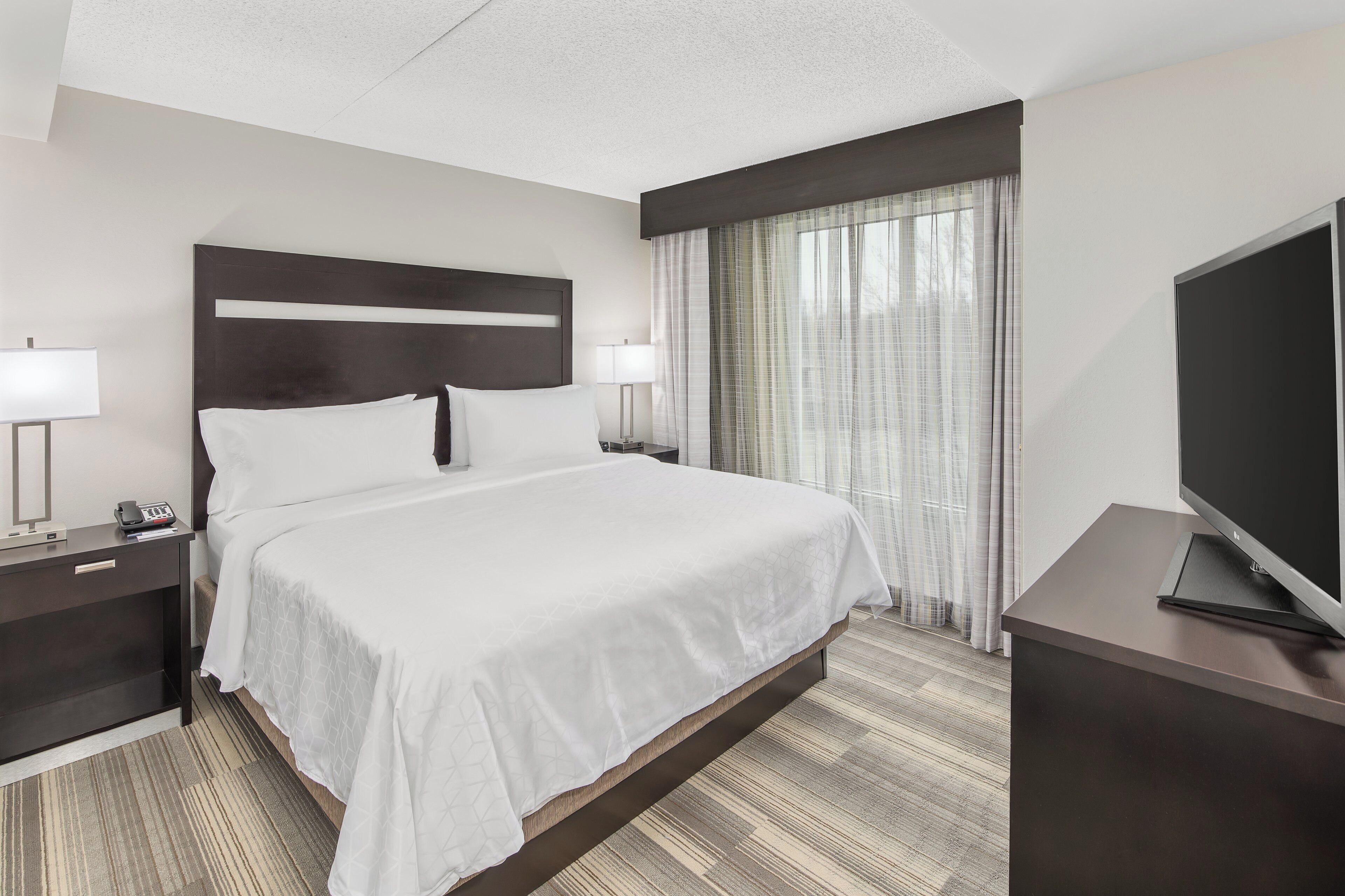 A hotel room with a large bed, two bedside tables, a TV, and a window with curtains.