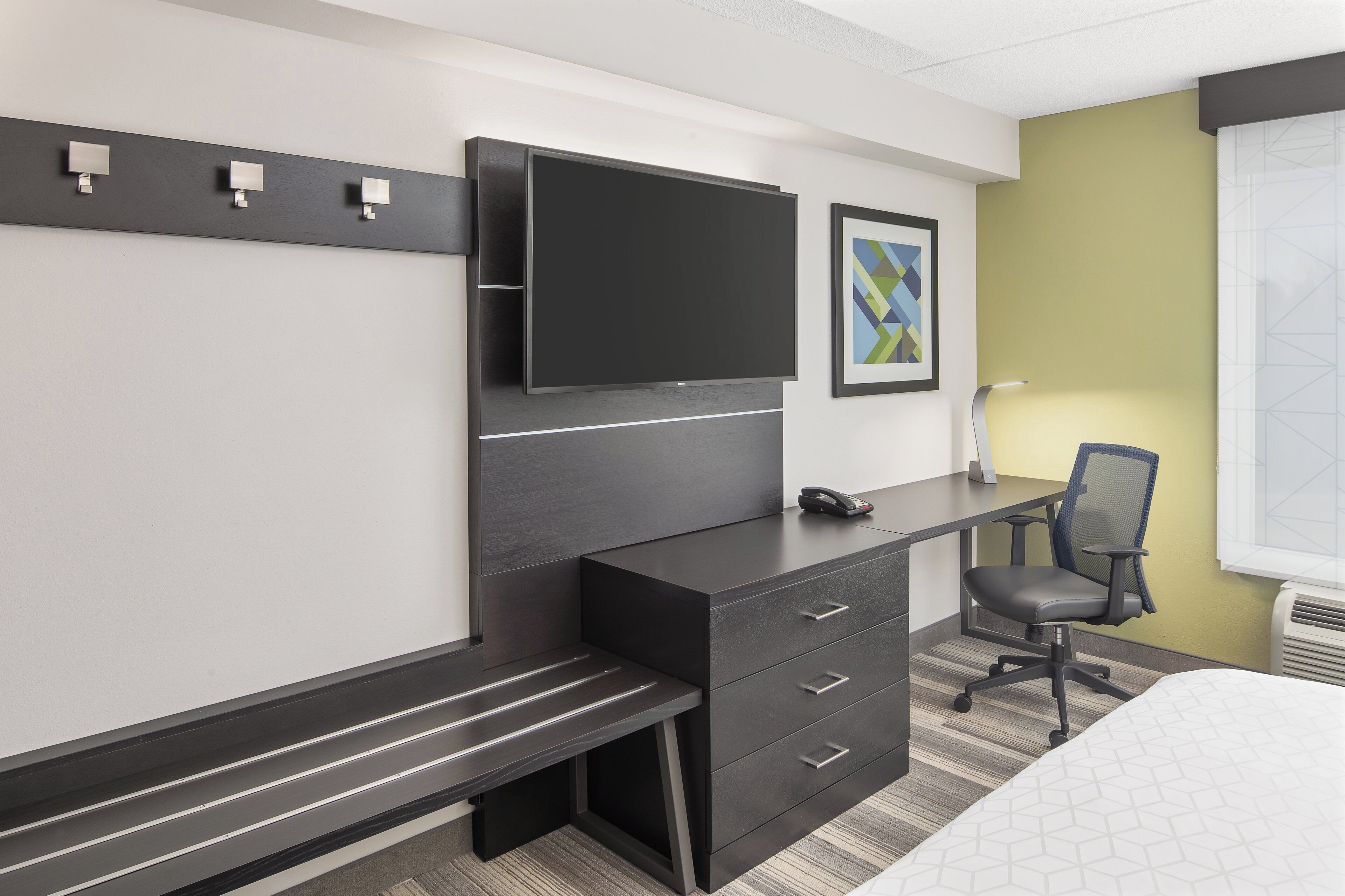 A hotel room with a bed, a desk with drawers, a chair, a flat-screen TV mounted on the wall, and a framed picture.