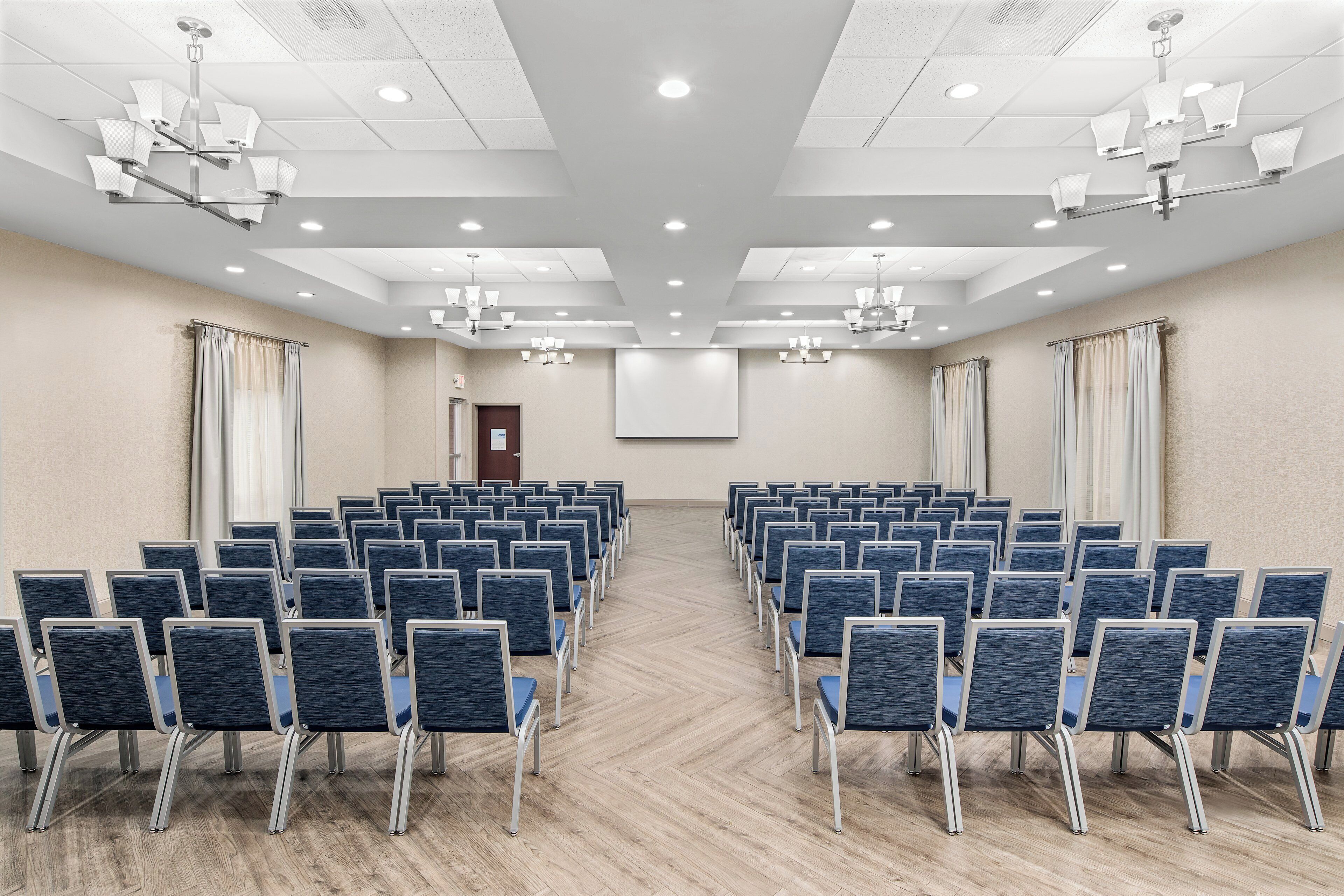Un salón de conferencias con filas de sillas, una pantalla de proyector y luces en el techo.