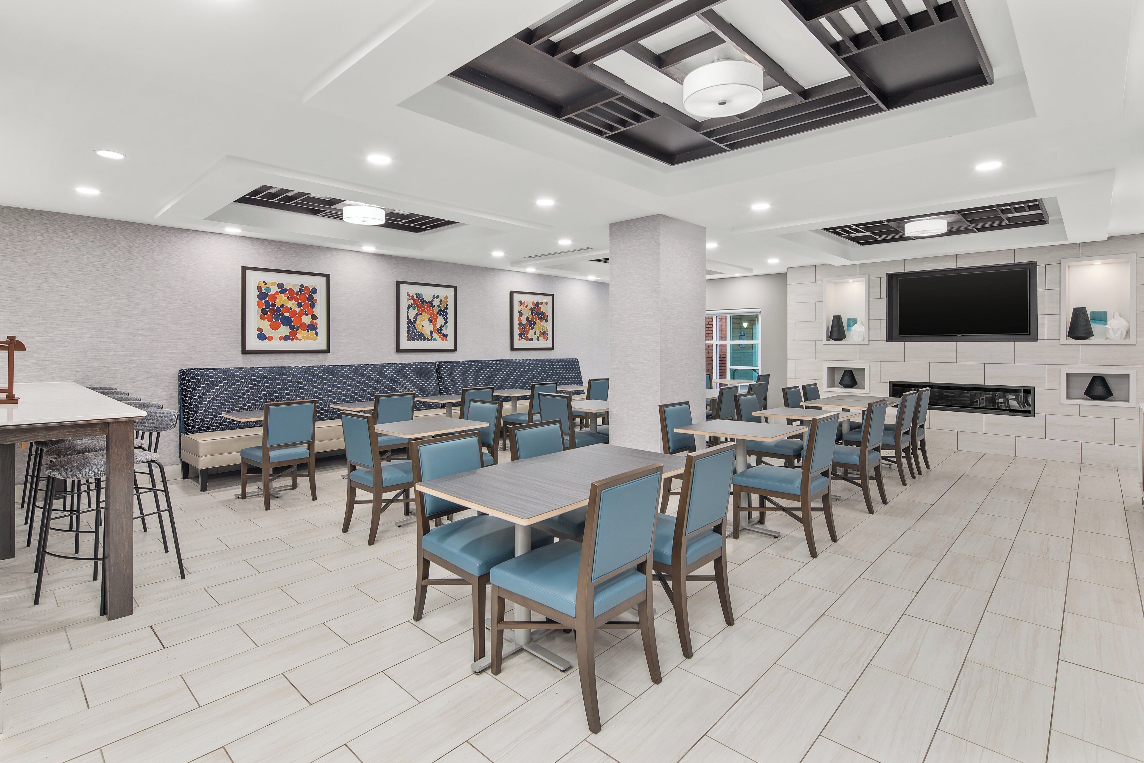 A modern restaurant with multiple dining tables and chairs, a seating area with a sofa, and a flat-screen TV mounted on the wall.
