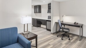 A modern hotel room with a blue sofa, a desk with a lamp, a chair, and a kitchenette in the background. - Holiday Inn Express & Suites Greenville-I-85 & Woodruff Rd by IHG (Greenville)