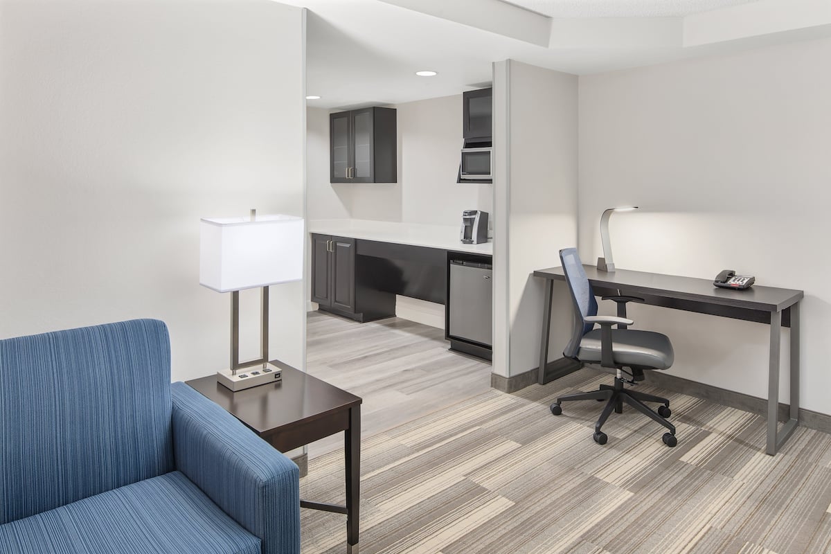 Une chambre d’hôtel moderne avec un canapé bleu, un bureau avec une lampe, une chaise et, au fond, une kitchenette.