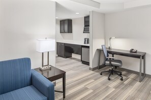 Een moderne hotelkamer met een blauwe bank, een bureau met lamp, een stoel en een kitchenette op de achtergrond.