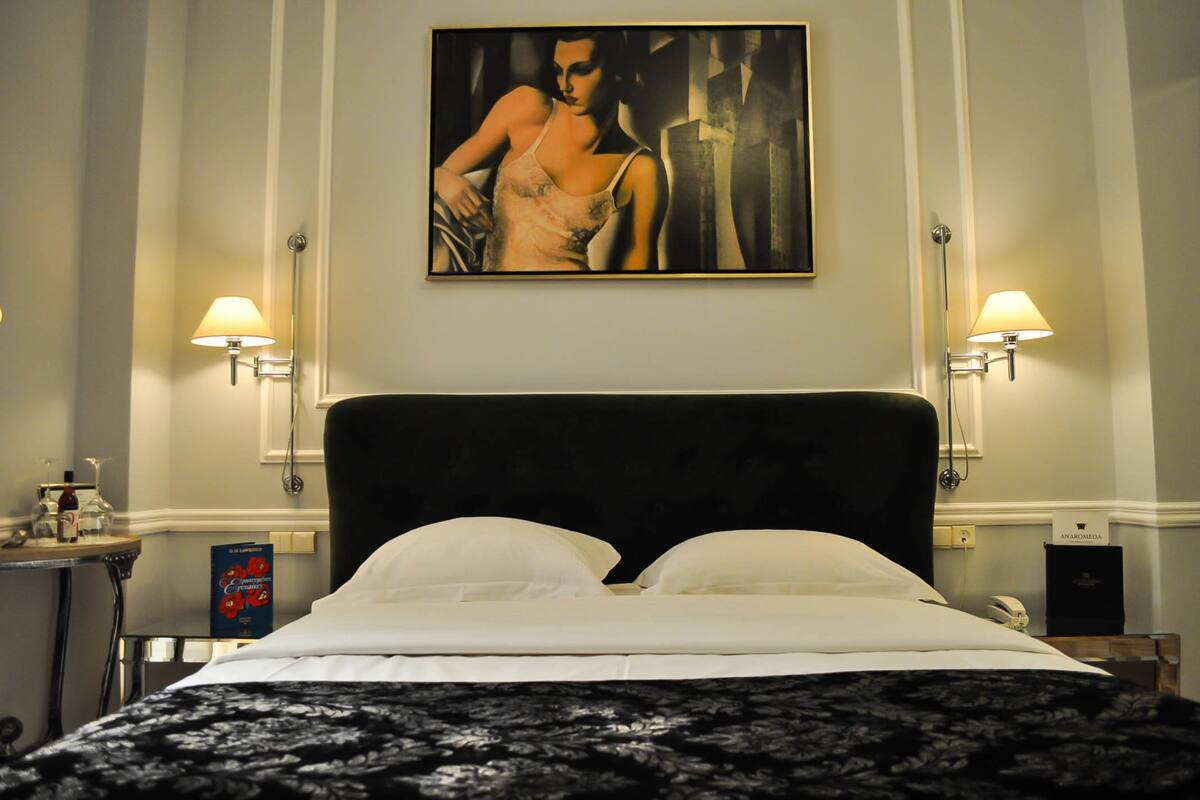 Habitació d'hotel amb un llit gran, dues làmpades de nit, un quadre a la paret i una taula petita amb un llibre i una ampolla.