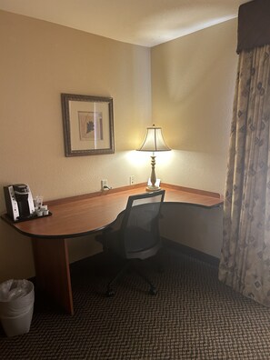 A hotel room with a wooden desk, a black office chair, a lamp, a framed picture on the wall, a coffee maker, and a trash bin. - Holiday Inn Express Las Vegas-Nellis by IHG (Las Vegas)
