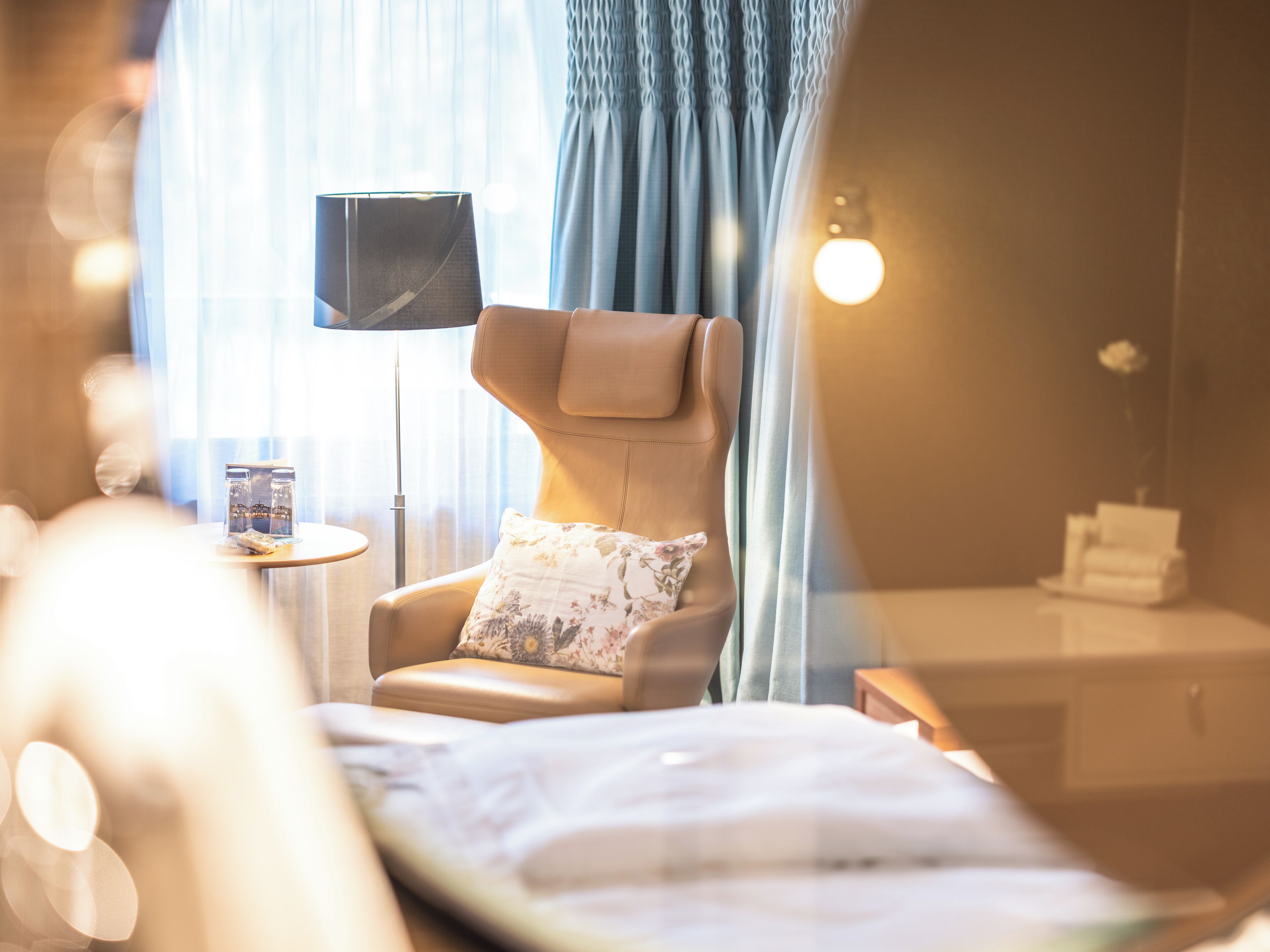Ein Hotelzimmer mit Bett, Sessel mit Kissen, kleinem Tisch mit Lampe und einem Fenster mit Vorhängen.