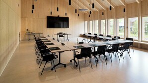 Un salón de conferencias con una larga mesa, sillas, una pantalla grande y luz natural proveniente de amplias ventanas.