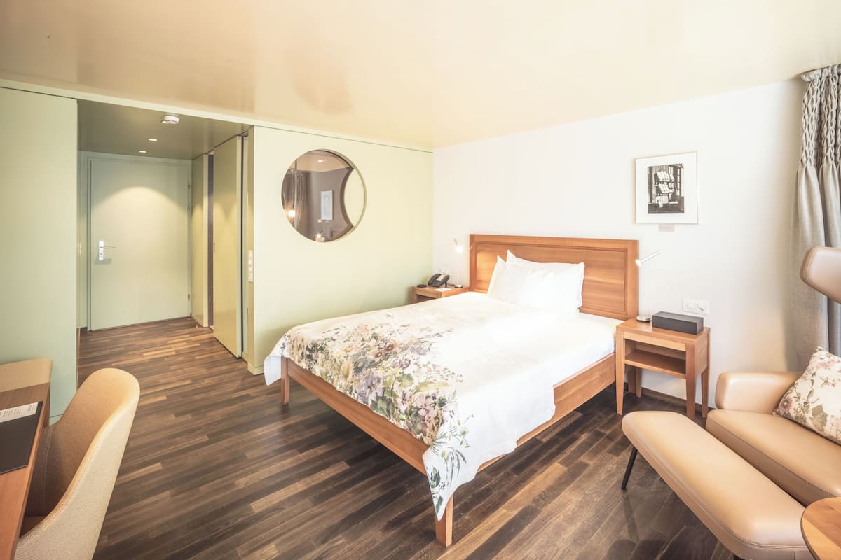 Une chambre d’hôtel avec un lit en bois, un miroir rond, une table de chevet avec une lampe, une chaise et un canapé.