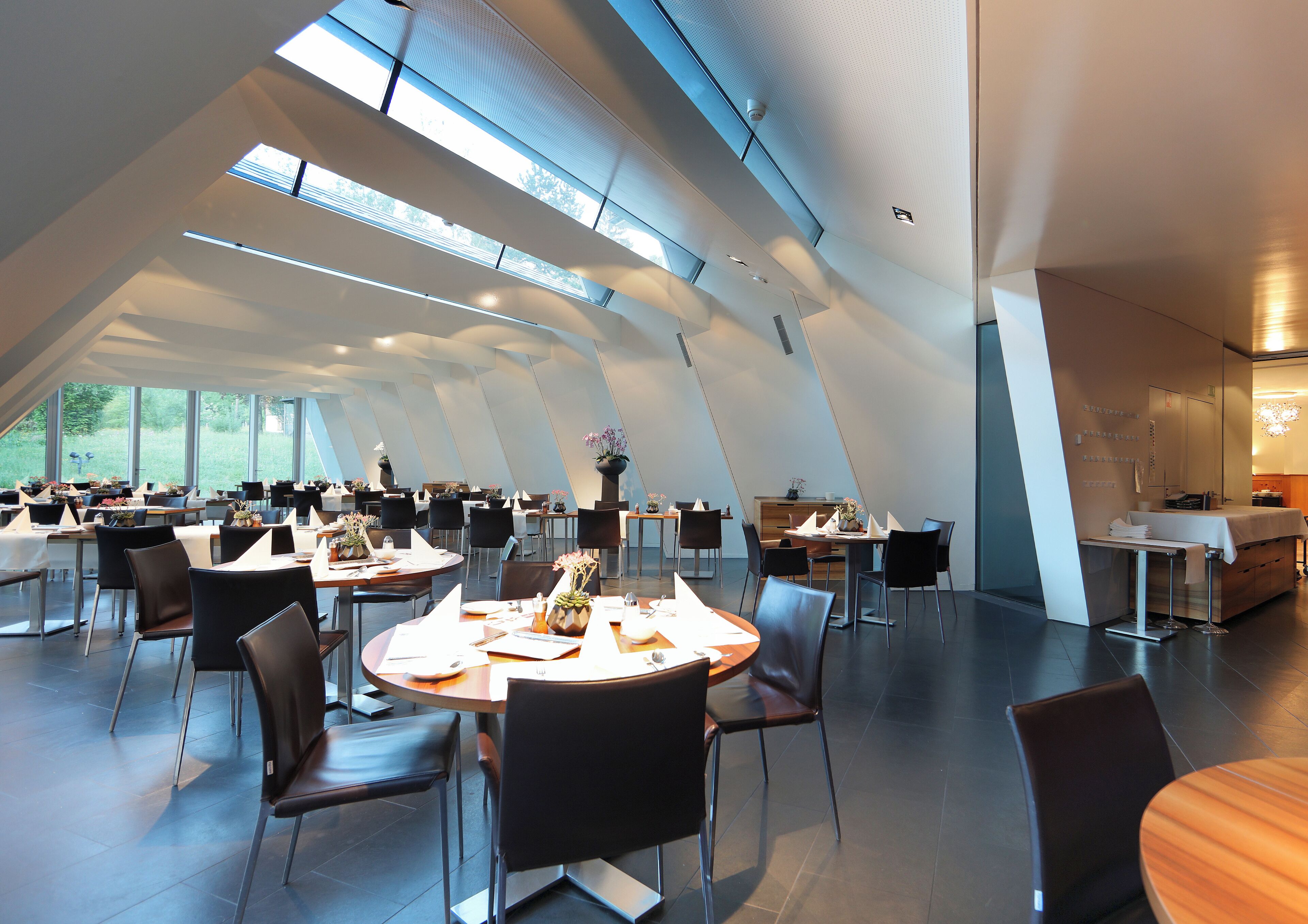 A modern dining area with multiple tables and chairs, large windows, and a high ceiling.